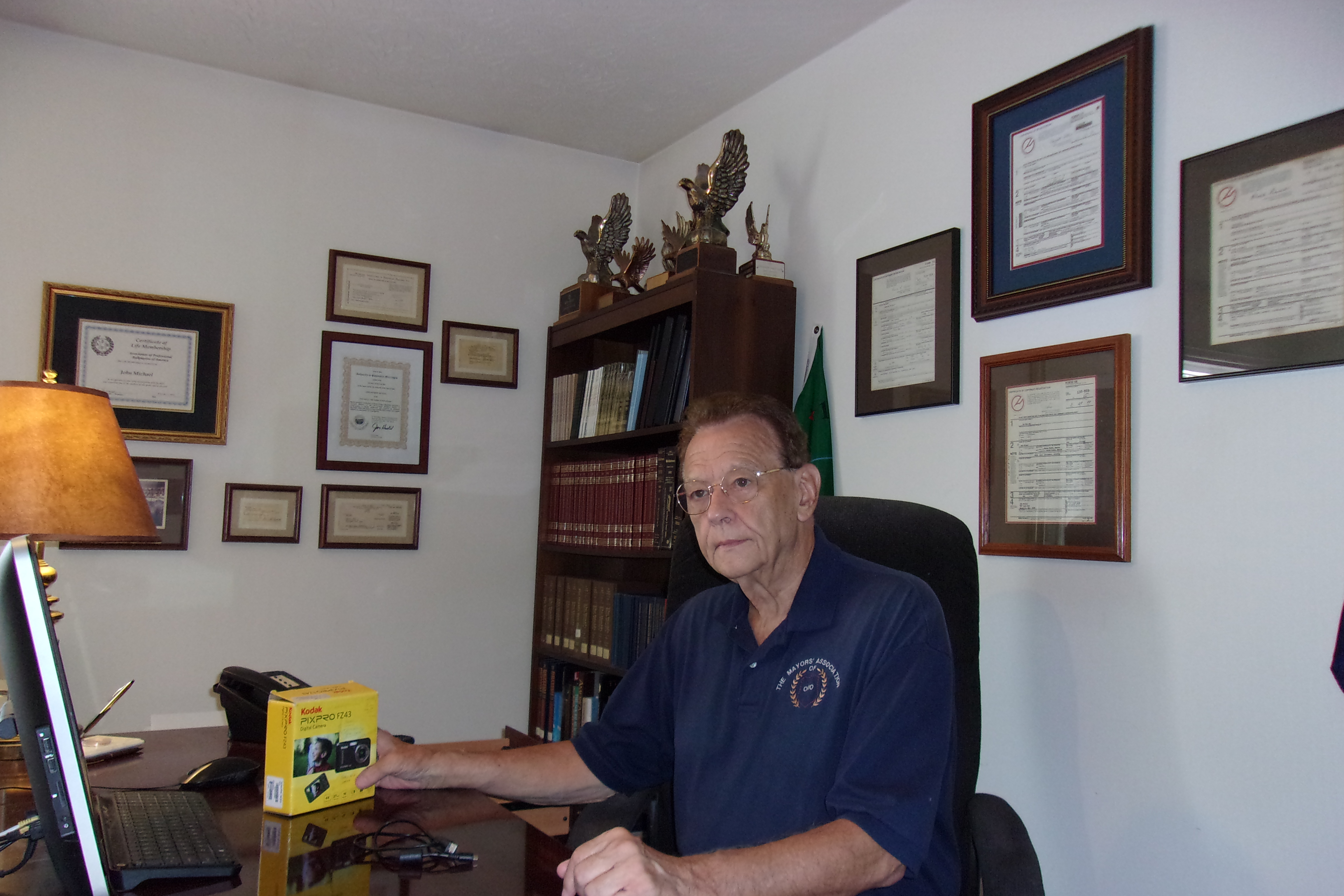 John Michael at his desk with credentials and awards on the wall behind him.